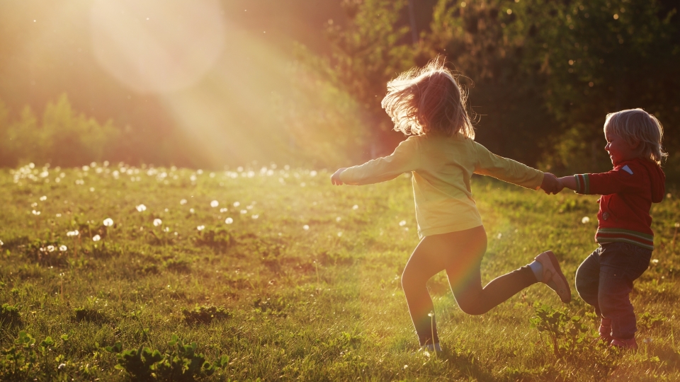 Zwei kleine Kinder laufen Hand in Hand über eine sonnendurchflutete Wiese, umgeben von Löwenzahn und warmem Abendlicht.